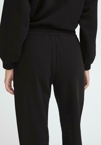 Melnas sporta bikses ar elastīgu jostasvietu. Materiāls izskatās gluds, cieši pieguļ jostasvietai un pakāpeniski sašaurinās līdz potītēm. Vienkāršs dizains.