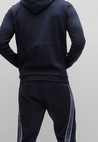 Mörkblå träningsoverall med huva, med en elegant design. Slät textur, kontrasterande vita sidostråk och en figurnära siluett.