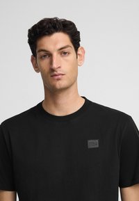 Schwarzes Baumwoll-T-Shirt mit Rundhalsausschnitt, das ein kleines schwarzes "POLO CLUB"-Patch auf der linken Brust trägt. Kurze Ärmel und glatte Textur.