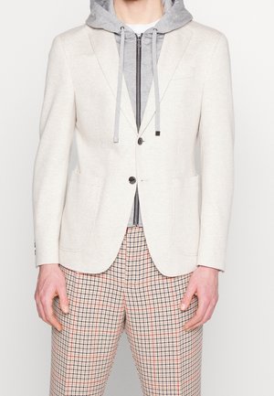 Mężczyzna w beżowej marynarce na szarej rozpinanej bluzie z kapturem i w kratkę spodniach, z rękami swobodnie opuszczonymi wzdłuż ciała na jednolitym tle.