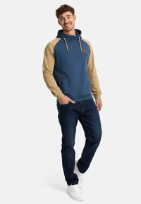 Sudadera con capucha azul y mangas color beige, que presenta un bolsillo frontal y cordones ajustables. Combinada con jeans oscuros y zapatillas blancas.