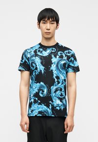 Άνδρας που φοράει μαύρο T-shirt με έντονα μπλε σχέδια κυμάτων ωκεανού, στέκεται μπροστά σε απλό λευκό φόντο.