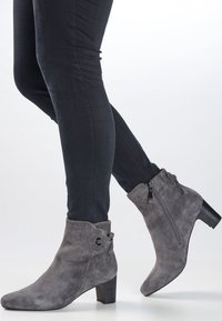 Harmaan mokkasiinivillainen nilkkuri, jossa on paksu korko, koristeellisia niittejä, sivuvetoketju ja nauhakoriste varressa.