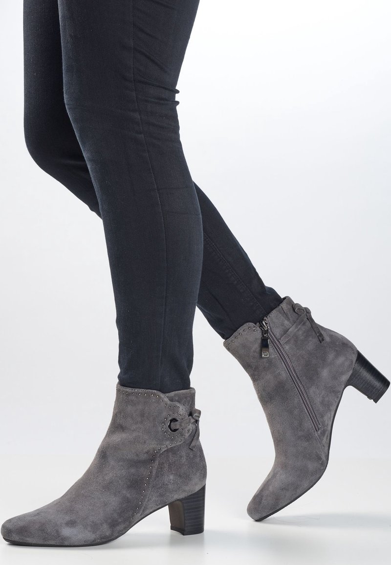 Harmaan mokkasiinivillainen nilkkuri, jossa on paksu korko, koristeellisia niittejä, sivuvetoketju ja nauhakoriste varressa.