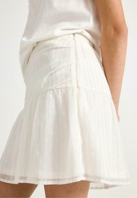 Hvid kort nederdel med vertikale stribemønstre og små, hævede prikkemønstre. Har en flared kant og samlet talje for tekstur.