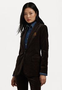 Καφέ σακάκι από κορντούρα με τακτοποιημένη εφαρμογή, διαθέτοντας δύο μπροστινές τσέπες και κεντημένο έμβλημα στο στήθος, φορεμένο πάνω από πουκάμισο τζιν.