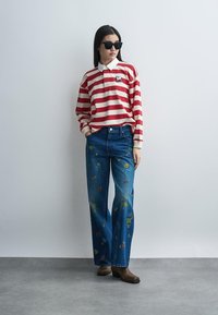 Nő fekete napszemüvegben, piros-fehér csíkos, hosszú ujjú inget és kék, virágos hímzésű farmert visel barna csizmával.