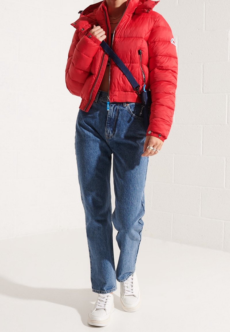 Doudoune rouge avec capuche, de longueur courte et fermeture éclair. Associée à un jean bleu taille haute et des baskets blanches. Comprend une sangle bleue.