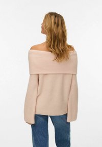 Frau mit welligem blonden Haar, die einen schulterfreien hellrosa Pullover und blaue Jeans trägt, mit dem Rücken zu einer schlichten weißen Wand.