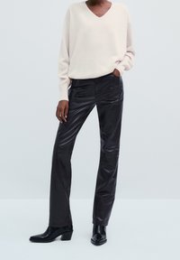 Person in einem cremefarbenen V-Ausschnitt-Pullover, schwarzen Lederhosen und schwarzen hohen Stiefeln, steht mit einer Hand in der Tasche.