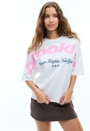 Junge Frau mit langen, welligen Haaren trägt ein weißes Oversize-T-Shirt mit dem Schriftzug „Brooklyn Crown Heights, New York U.S.A“ und einen dunklen Minirock.