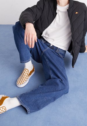 Osoba siedząca na niebieskiej podłodze, ubrana w czarną pikowaną kurtkę, białą koszulę, szerokie niebieskie dżinsy, białe skarpetki oraz brązowo-białe slipony w kratkę.