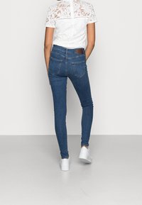 Jeans de denim azul ajustados con cintura alta, bolsillos traseros y un parche de cuero marrón en la cintura, combinados con una blusa blanca de encaje.