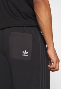 Svarta fleecebyxor har en sidoficka med vit Adidas-logga. Tre mörka ränder löper ner för utsidan av benet. Slät textur.