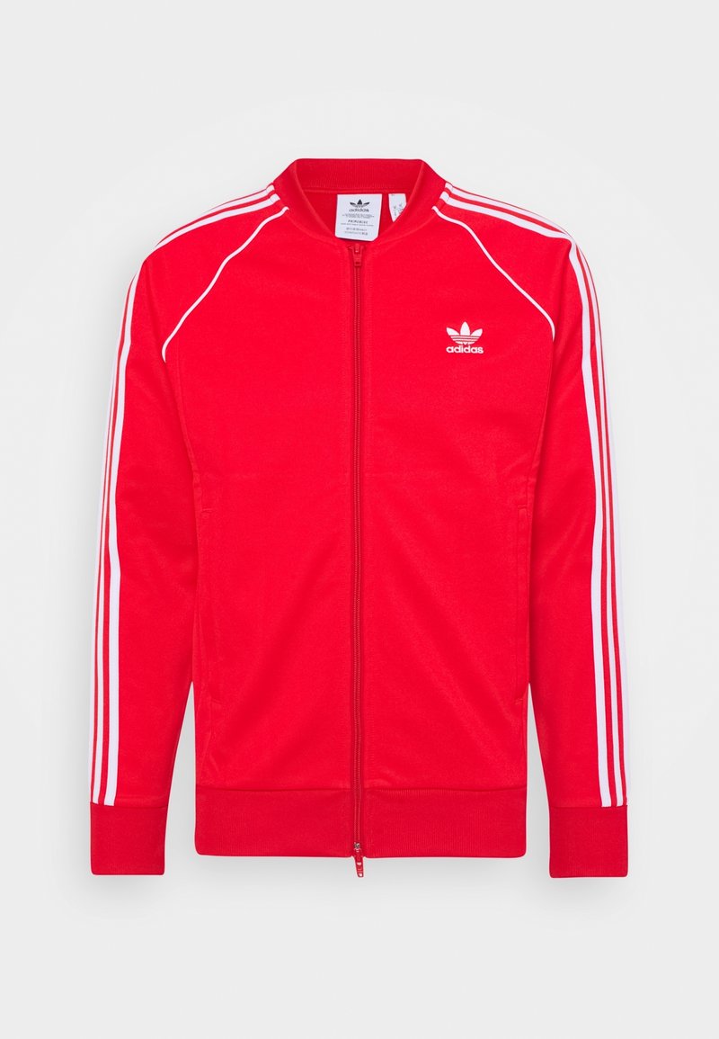 Červená bunda Adidas s bílými pruhy na rukávech, předním zipem, žebrovanými manžetami a logem Adidas trefoil na levé straně hrudi.
