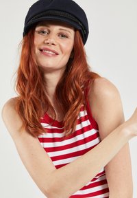 Rood-wit gestreepte tanktop met een ribbelstructuur, gecombineerd met een donkerblauwe pet met een gevlochten accent. Persoon glimlacht, met ontspannen armen.