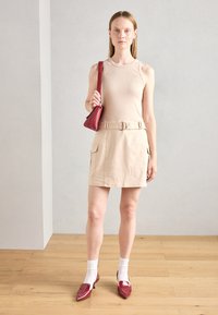 Débardeur beige à côtes, jupe beige avec ceinture, sac à main rouge et chaussures à talons rouges brillantes avec des chaussettes blanches. Fond uni et sol en bois.