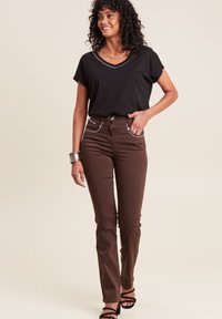 Schwarzes V-Ausschnitt-T-Shirt kombiniert mit hochgeschnittenen braunen Hosen, die weiße Nähte an den Taschen aufweisen. Das Model trägt schwarze Riemchenschuhe und silberne Accessoires.