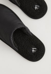 Chaussons noirs avec une surface matelassée, une doublure intérieure en polaire noire douce, une semelle texturée et une petite étiquette logo sur la partie supérieure.