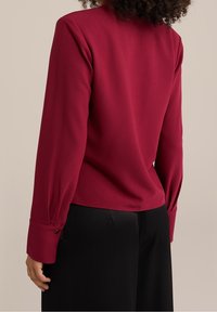 Blusa rossa a maniche lunghe con collo alto e polsini con bottoni, abbinata a pantaloni neri plissettati, caratterizzata da una texture liscia e morbida.