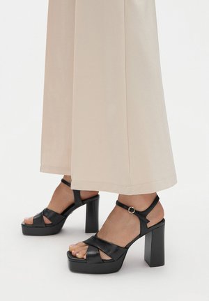 Sandalias de plataforma negras con gruesos tacones cuadrados, con correas cruzadas y una delgada correa en el tobillo con un acento metálico. Textura de cuero suave.