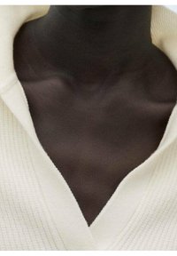 Weißer gerippter Strickstoff bildet einen tiefen V-Ausschnitt, der im Kontrast zur glatten, dunklen Haut steht. Der Fokus liegt auf der Textur und den Stoffdetails.