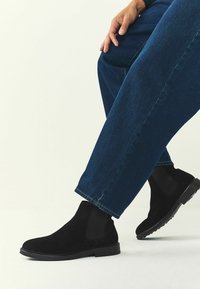 Mustade sametist ankle-boots, millel on elastsed külg-paneelid, sile tald ja ümar varvas, koos siniste denimipükstega.
