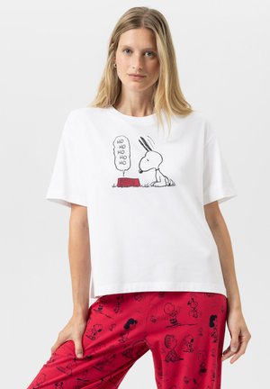 Blonde Frau trägt ein weißes T-Shirt mit Snoopy und dem Text "Ho Ho Ho" über einer roten Hundeschüssel, kombiniert mit roten Hosen, die Peanuts-Charaktere zeigen.
