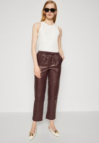 Haut sans manches blanc, pantalon en cuir bordeaux avec taille à cordon, et chaussures blanches à bout pointu avec accents dorés. Texture lisse sur l'ensemble.