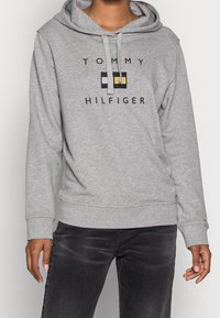 Szary hoodie wykonany z miękkiego materiału z kapturem na sznurku. Posiada logo "TOMMY HILFIGER" oraz prostokątny wzór w kolorze granatowym, białym i żółtym.