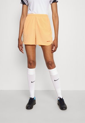Person trägt hellorange Nike FC Sportshorts, weiße Nike knielange Socken, schwarze Fußballschuhe und ein weißes Sportshirt und steht vor einem neutralen Hintergrund.