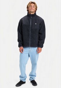 Fliisist jakk tumesinise kehaga ja heledama nailonist ülaosaga. Omaduseks on kõrge krae, tõmblukuga sulgur ja bränditud rinnatasku.