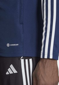 Tumesinine spordisärk tekstuuriga kangast, millel on valged kolmestriibud varrukatel ja AeroReady logo allääres. Allpool on mustad püksid.