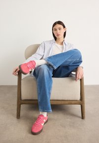 Rosa suedetrenere med snøring, hvite detaljer, brukt med løse blå jeans og en hvit t-skjorte, kombinert med en stripete skjorte, sittende på en lys stol.