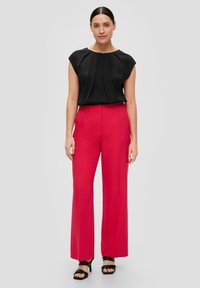 Top nero a maniche corte con pieghe, abbinato a pantaloni rossi a vita alta e gamba larga. Scarpa nera con cinturini completa l'outfit.
