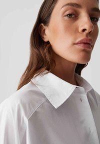 Jonge vrouw met lang bruin haar, gekleed in een wit overhemd met kraag, die zelfverzekerd opzij kijkt tegen een neutrale achtergrond.