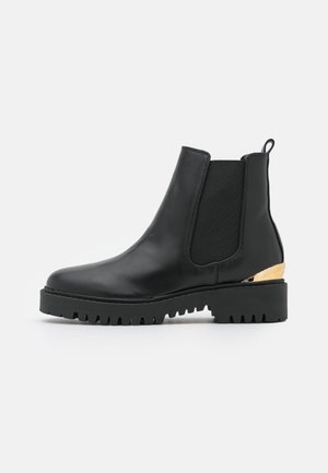 Botte Chelsea en cuir noir avec panneaux latéraux élastiques, semelle crantée épaisse, languette à l'arrière et accent métallique doré sur le contrefort du talon.