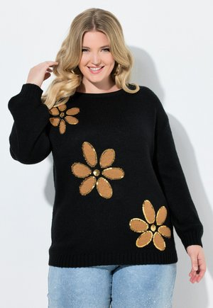 Mujer rubia sonriendo, con un suéter negro con tres grandes flores marrones y doradas con lentejuelas y vaqueros azul claro.