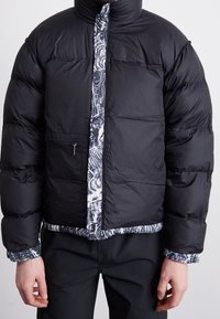 Sort dunjakke med en skinnende finish, fremtrædende quiltede sektioner og en mønstret blå og hvid lynlåsforside.
