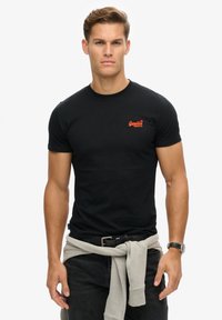 T-shirt en coton noir à manches courtes, avec un logo orange sur la poitrine. Assorti avec un jean noir et un sweat gris noué à la taille.