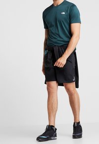 Camiseta atlética de manga corta color verde azulado con logo, pantalones cortos deportivos negros con bolsillos y zapatos negros con detalles reflectantes; atuendo casual para realizar actividad.