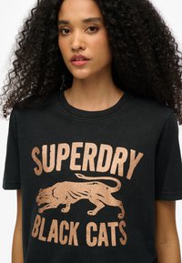 Camiseta de algodón negra con gráficos metálicos en color oro rosa de las palabras "SUPERDRY" y "GATOS NEGROS" con un diseño de león. Cuello redondo, ajuste relajado.