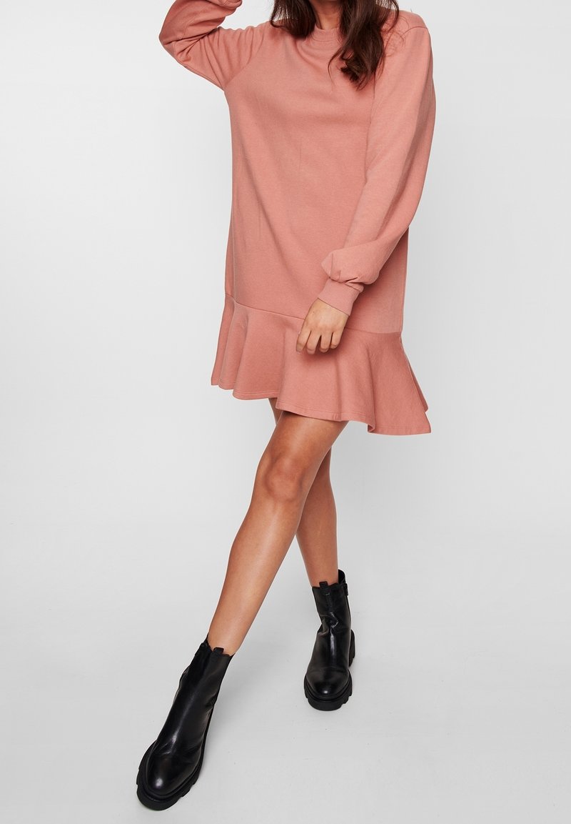 Robe rose à manches longues avec un ourlet évasé, confectionnée dans un matériau doux. Associée à des bottines noires à semelle épaisse.