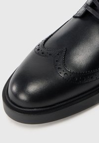 Pantof de piele neagră tip brogue cu perforații decorative și talpă groasă din cauciuc, prezentat în prim-plan pe un fundal alb.