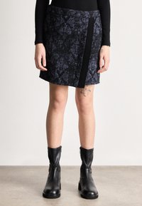 Femme portant une jupe à motifs noirs et bleu foncé, un haut noir à manches longues, des bottes en cuir noires mi-mollet, dévoilant des tatouages sur ses jambes.