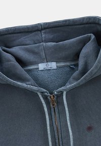 Hoodie zippé gris avec un intérieur doux, doté d'une fermeture éclair en bronze et de coutures contrastantes. Étiquette à l'intérieur portant le nom de la marque.