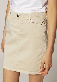 Gonna mini in denim beige con tasche, passanti per la cintura, chiusura frontale con bottoni e dettagli di cucitura sottili. Tessuto liscio con una vestibilità su misura.