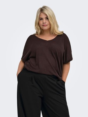 Femme blonde portant un haut ample à manches courtes marron foncé et un pantalon noir, les mains dans les poches, debout devant un fond clair uni.