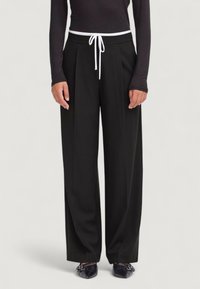 Pantaloni neri a gamba larga con una cintura elastica bianca, caratterizzati da pieghe e una texture liscia, abbinati a scarpe nere.