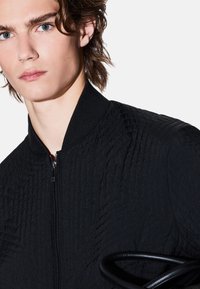 Giacca bomber nera con un motivo testurizzato, chiusura a zip frontale e colletto a coste. Il modello tiene un accessorio con un manico circolare nero.